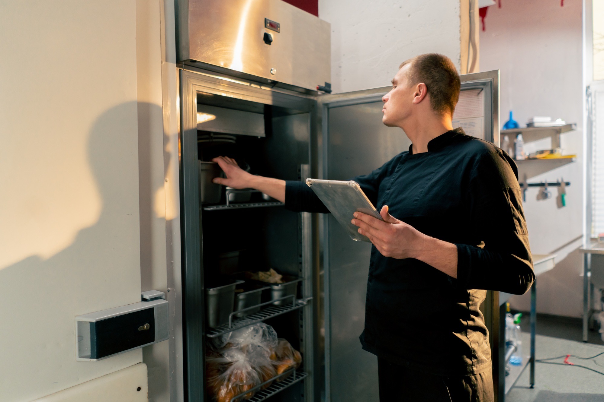 chef stands in kitchen in a black uniform and looks through what is on the list in the refrigerator