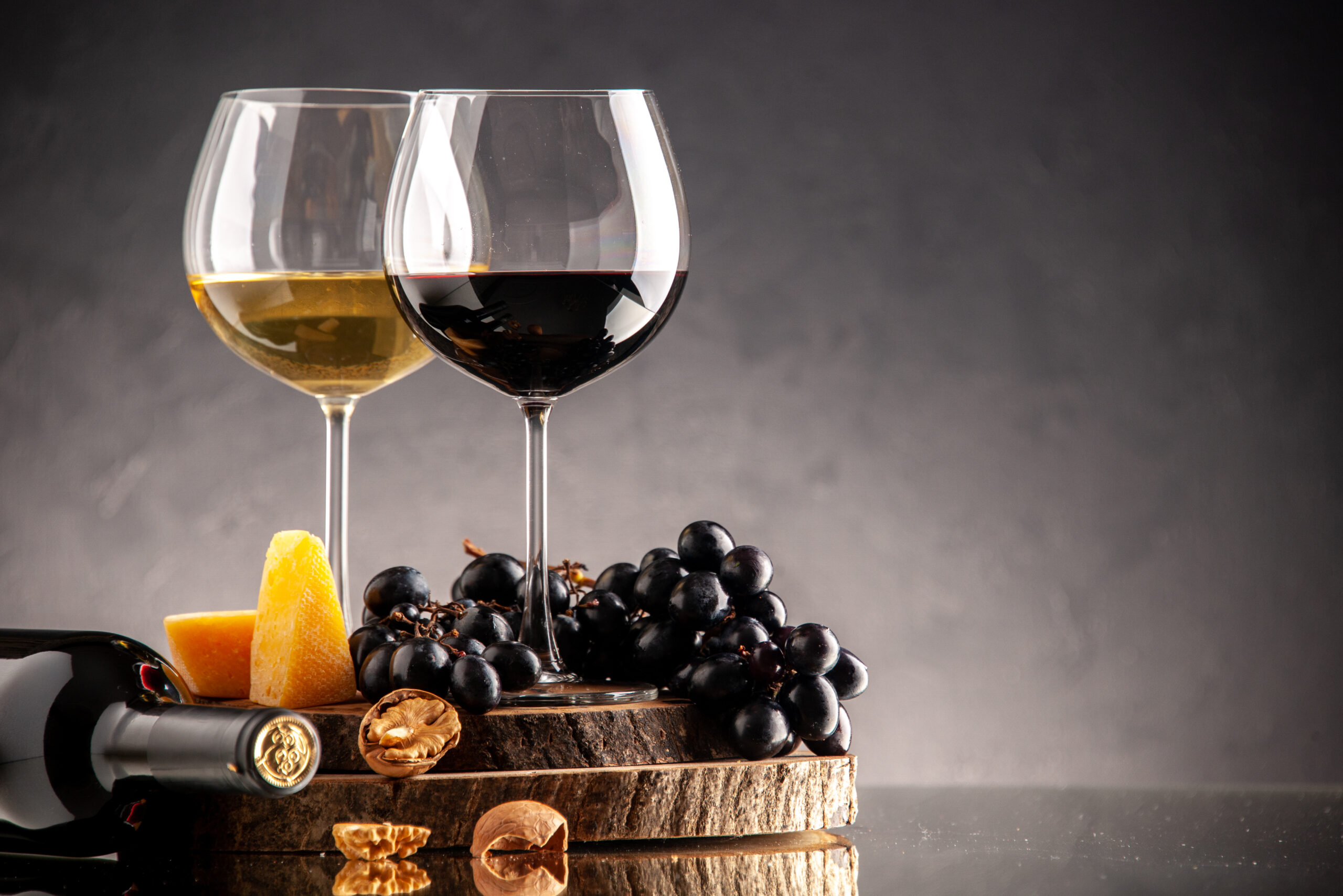 vista-frontal-copas-vino-uvas-frescas-nueces-queso-amarillo-sobre-tablero-madera-botella-volcada-sobre-fondo-oscuro